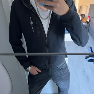 Marinblå zip hoodie från sail racing - Snygg marinblå zip hoodie från Sail Racing med dragkedja framtill och logga på bröstet. Jackan har huva med snörning och ribbade muddar. Perfekt för en chill och sportig look, funkar lika bra till jeans som till träningsbyxor.