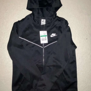 Nike Tape ofta - Nike Tape Kofta – Svart (Unisex) Stilren och sportig Nike Tape zip-hoodie i svart med klassiska logoband längs ärmarna. Perfekt för både vardag och träning. Mjuk bomullsblandning gör den bekväm att bära hela dagen.