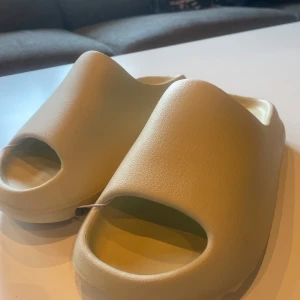 Beige slides från Adidas - Chunky beige slides från Adidas med öppen tå och platt sula. Tillverkade i syntetmaterial med minimalistisk design och diskret Adidas-logga på innersulan. Perfekta för chill dagar eller att glida runt i hemma eller på stranden.