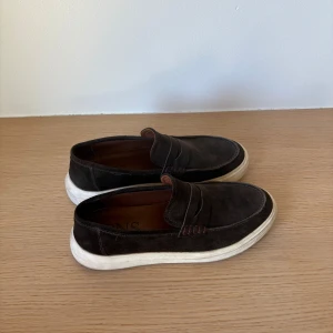 Bruna mockaloafers från CNS - Snygga loafers i mörkbrun mocka från CNS med klassisk pennyloafers-design och kontrasterande vit sula. Skorna har rund tå och diskreta sömdetaljer, perfekta för en chill och stilren look.