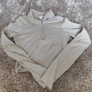 Beige långärmad tröja från Monki - Säljer en beige långärmad tröja från Monki i mjuk bomull. Tröjan har en klassisk krage och en half zip-dragkedja framtill som ger en chill och stilren look. Perfekt att matcha med jeans eller kjol för en enkel och snygg outfit.