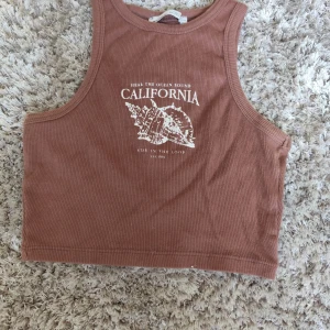 Brun ribbad linnetopp Pull&Bear XS - Säljer en brun ribbad linnetopp från Pull&Bear i storlek XS. Toppen har ett tryck med snäcka och texten 'California' framtill. Croppad modell med ärmlös design, perfekt för varma dagar och chill vibes.