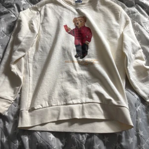 Vit sweatshirt Polo Bear Ralph Lauren s - Vit långärmad sweatshirt från Polo Ralph Lauren i bomull, med ikoniska Polo Bear-trycket framtill där björnen har röd jacka och håller ett kort. Klassisk rund halsringning och relaxed fit. Perfekt för dig som stilrena plagg.  Passar folk i S, köpt i kids brandstore ( själv är jag är 178 och väger 65 kg och passar utmärkt.