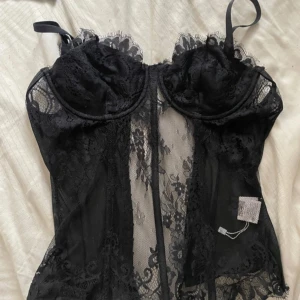 Svart spetsig korsett med tunna axelband - Säljer en svart korsett med tunna justerbara axelband och vacker spetsdetalj över hela toppen. Den har genomskinliga partier och bygel vid bysten för extra form. Perfekt för dig som vill ha en edgy och snygg look.