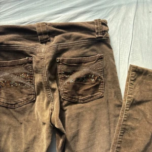 Bruna manchesterbyxor med strassdetaljer - Bruna manchesterbyxor från Authentic med snygga strassdetaljer på bakfickorna och en unik knapp framtill med bruna stenar. Byxorna har normal passform och raka ben. Perfekta för dig som vill ha något extra i din outfit. Byxorna är INTE skinny utan raka, alltså raka ben. Byxorna saknar några stenar på framsidan och på fickan, men det är inget som stör funktionen och looken av de