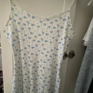 Vit blommig sommarklänning med smala band - Supersöt vit klänning med små blå blommor och smala axelband. Lätt ribbat tyg och vågig kant upptill ger en gullig vibe. Perfekt för varma dagar och enkel att styla med sneakers eller sandaler.