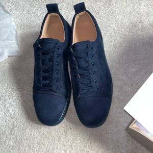 Mörkblå sneakers från Louboutin - Snygga mörkblå sneakers från Christian Louboutin i mocka med klassisk röd sula och rund tå. Skorna har snörning och en stilren design som sticker ut med den ikoniska röda undersidan. Perfekta för dig som vill ha något exklusivt och trendigt.