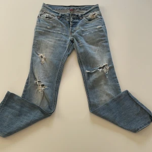 Ljusblå lågmijade bootcut jeans - Säljer ett par ljusblå jeans med bootcut passform och snygga slitningar och hål på benen. Jeansen har klassisk femficksdesign, låg midja och är tillverkade i mjukt denim. Perfekta för dig som gillar en avslappnad och trendig look.