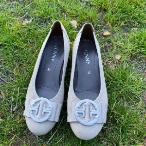 Beige ballerinaskor från Jenny, strl 37 - Snygga beige ballerinaskor från Jenny i mocka-look med rund tå och låg klack. Framtill sitter en dekorativ rosett och ett stort runt emblem i ljusgrått. Perfekta för dig som gillar klassisk och stilren design.