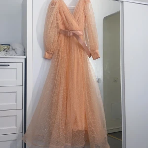 Lång klänning dammrosa färg  - Drömmig långklänning i persikoton från Dekolte med transparent tyll, små prickar och v-ringad omlottfront. Puffiga långärmar och tillhörande bälte i midjan. Perfekt för dig som vill sticka ut med en romantisk och elegant look.