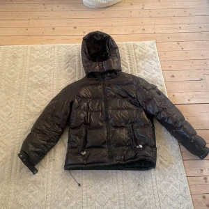 Moncler Fashion puffer jacka  - Säljer en svart pufferjacka med glansig finish och huva. Jackan har dragkedja framtill, två fickor med dragkedja och justerbar nederkant med snörning. Perfekt för kalla dagar när du vill hålla stilen och värmen.