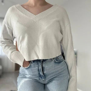 Vit croppad stickad tröja med v-ringning - Snygg vit croppad stickad tröja med bred ribbad kant och v-ringad hals. Tröjan har långa ärmar med ribbade muddar och en avslappnad passform. Perfekt att matcha med höga jeans för en trendig look.