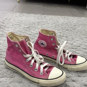 Rosa Converse All Star high tops - Snygga rosa Converse All Star high tops med klassisk vit gummitå och vita snören. Skorna har den ikoniska loggan på sidan och svart rand längs sulan. Tillverkade i canvas med platt sula och rund tå. Perfekta för dig som vill sticka ut med färg!