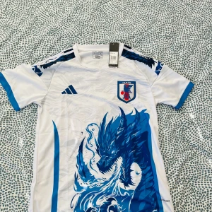 Japan Adidas fotbollströja drakmotiv - Japan landslagets fotbollströja från Adidas i vitt med blå detaljer och ett stort blått drakmotiv på framsidan. Tröjan har korta ärmar, rund hals och klubbmärke på bröstet. Tillverkad i lätt och ventilerande material, perfekt för match eller träning.