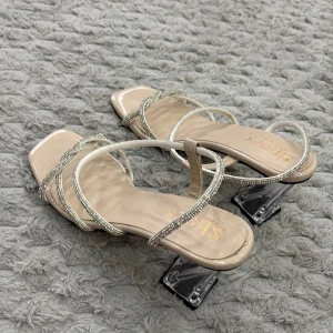 Beiga sandaler med strass och klack - Snygga beiga sandaler med fyrkantig tå, tunna remmar dekorerade med glittrande strass och unik transparent klack. Perfekta för dig som vill sticka ut med en elegant och trendig look. Tillverkade i syntetmaterial.