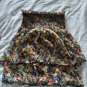 Blommig kortklänning med volang - Supersöt kjol med smock upptill och volangig kjol. Klänningen har ett färgglatt blommönster i rosa, blått, gult och grönt på en mörk botten. Perfekt för sommarens alla tillfällen och ger en riktigt romantisk vibe.