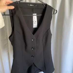 Svart skräddarsydd väst Gina Tricot - Snygg svart väst från Gina Tricot med skräddad passform och v-ringning. Västen har tre knappar framtill och är ärmlös med klassisk look. Oanvänd med originalpris på 459 kronor 