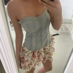 Axelbandslös klänning med jeansliv - Supersöt axelbandslös klänning med ljusblått jeansliv och volangig kjol i blommigt mönster. Kjolen är kort och har en romantisk vibe med rosa och röda blommor på vit botten. Perfekt för sommarens alla tillfällen!