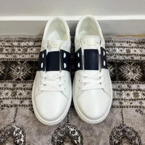 Valentino Garavani Open sneakers vit/svart - Snygga vita Valentino Garavani Open sneakers med bred svart rem över vristen och coola nitar på hälen. Skorna har rund tå, vita snören och är gjorda i skinn. Perfekta för dig som vill ha en stilren men ändå edgy look.