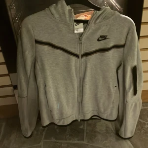 Grå Nike hoodiejacka med dragkedja - Snygg grå hoodiejacka från Nike med svart logga på bröstet och svarta detaljer längs bröstet och ärmarna. Jackan har hel dragkedja framtill, huva och en cool ficka med dragkedja på ärmen. Perfekt för dig som gillar sportig och avslappnad stil.