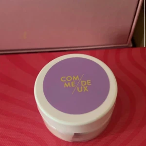 Comme Deux aha exfoliating  - Highlighter från Comme Deux i en rund vit plastburk med lila lock och gul text. Perfekt för att ge glow till ansiktet och framhäva kindbenen. Smidig att ta med i väskan och enkel att applicera med fingertoppar eller borste.