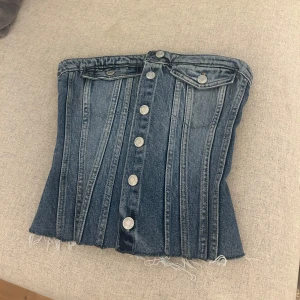 Jeanskorsett med knappar från Zara - Säljer en snygg jeanskorsett från Zara i klassisk blå denim. Toppen är axelbandslös med silverfärgade knappar framtill. Bra skick endast använd enstaka gånger.🩵