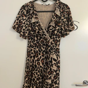 Leopardmönstrad omlottklänning med volang från Boohoo - Kort leopardmönstrad klänning med omlott och volangdetaljer längs urringningen och framtill från Boohoo i storlek 34. Klänningen har korta, vida ärmar och v-ringad hals. Finns knytband till midjan. Klänningen är i mycket bra skick.