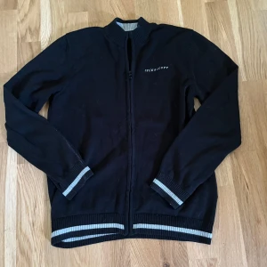 Svart kofta från Lyle & Scott med dragkedja - Svart kofta från Lyle & Scott med hel dragkedja och ribbade muddar i svart och grått. Broderad gul logga på ärmen och diskret Lyle & Scott-tryck på bröstet. Perfekt för dig som gillar klassisk stil med sportig touch.