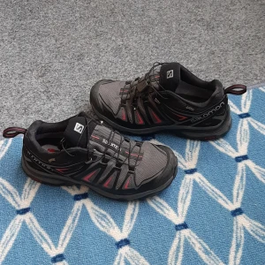 Salomon svarta sneakers med röd detalj - Grymma svarta Salomon sneakers med röda och grå detaljer. Skorna har snörning med snabbsnörningssystem, robust yttersula och meshpaneler för extra andningsförmåga. Perfekta för äventyr och vardag, med cool outdoor-vibe och snygg design.