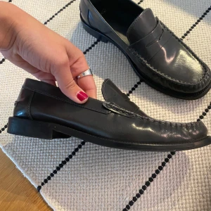 Svarta loafers i från MNG nya - Klassiska svarta loafers från MNG i glansigt skinn. Snygg penny loafer-design med markerade sömmar runt tån och platt sula. Perfekta för en clean och stilren look, passar till både jeans och kostymbyxor eller till en coolare outfit.