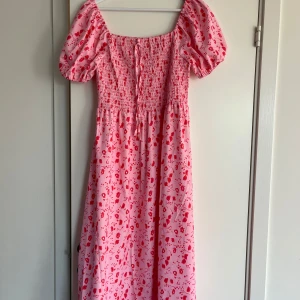 Rosa långklänning med puffärm L - Supersöt rosa långklänning med röda blommor, puffärmar och smockad överdel. Klänningen har knytband framtill och en luftig, flowy kjol. Perfekt för sommarens alla tillfällen när du vill känna dig extra fin.