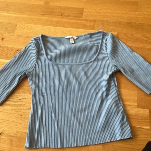 Ljusblå ribbad topp från H&M - Supersnygg ljusblå ribbad topp från H&M med fyrkantig ringning och trekvartsärm. Perfekt att styla med jeans eller kjol för en chill look. Mjuk och stretchig i materialet för extra komfort.
