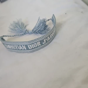 Dior armband  - Supersnyggt blått armband från Christian Dior med texten 'J'Adior' broderad i vitt. Armbandet är gjort i bomull och har dekorativa tofsar i ändarna för en trendig look. Perfekt accessoar för att lyfta din outfit.