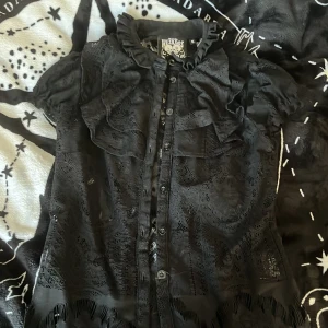 Svart spetsblus Killstar S - Supersnygg svart blus i spets från Killstar. Blusen har volangdetaljer vid axlar och krage, knappar framtill och korta ärmar. Genombrutet spetsmönster ger en edgy vibe och den höga kragen gör den extra unik. Perfekt för dig som gillar alternativ stil.