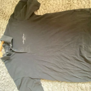 Svart pikétröja H&M Paris-brodyr - Svart pikétröja från H&M med klassisk krage och tre knappar framtill. På bröstet finns en broderad text med Paris-inspiration. Tröjan är i bomull och har korta ärmar, perfekt för en clean och stilren look.