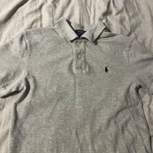 Grå pikétröja från Polo Ralph Lauren - Klassisk grå pikétröja från Polo Ralph Lauren med korta ärmar och krage. Tröjan har två knappar framtill och den ikoniska broderade loggan i mörkblått på bröstet. Perfekt basic till garderoben och funkar året runt.