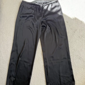 Svarta vida byxor från BikBok - Snygga svarta byxor från BikBok i storlek L. De har en mjuk, glansig yta och resår i midjan med snörning. 