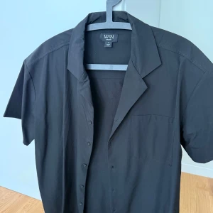 Svart kortärmad skjorta BoohooMAN - Svart skjorta från BoohooMAN i storlek M. Skjortan har klassisk krage, knäppning framtill och en bröstficka. Enkel och stilren design som passar perfekt till jeans eller shorts. Sitter ganska oversize så passar även de som bär storlek L skulle jag säga. Knappt använd! Tveka inte att hör av er vid frågor, mvh.