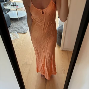 Persikofärgad långklänning med öppen rygg - Snygg långklänning i persikofärg från design by si, med smala axelband och öppen rygg med knyt. Klänningen har en mjuk satinliknande yta och en liten cut-out framtill. Perfekt för sommarens alla tillfällen. Aldrig använd 