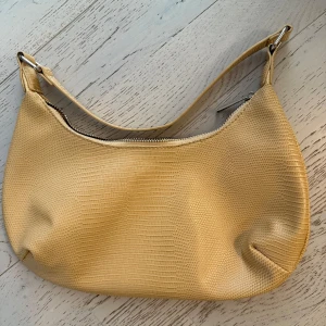 Gul axelväska med reptilmönster - Säljer en gul axelväska i syntetmaterial med reptilinspirerat mönster. Väskan har en mjuk, rundad form och stängs med en silvrig dragkedja. Perfekt storlek för det viktigaste och har en enkel, stilren look.