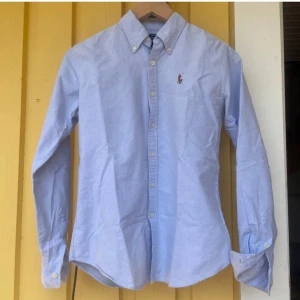 Ljusblå skjorta från Ralph Lauren - Klassisk ljusblå skjorta från Ralph Lauren i slim fit-modell. Skjortan har långa ärmar, knappar framtill och button down-krage. Broderad logga på bröstet. Tillverkad i bomull, perfekt för en clean och stilren look.