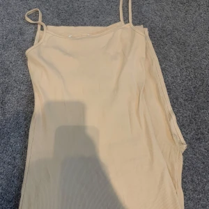 Beige långklänning med smala axelband - En stilren beige långklänning med smala axelband och figurnära passform. Klänningen är ärmlös och har en enkel, clean look som passar perfekt för varma dagar. Materialet är ribbat och känns mjukt mot huden.