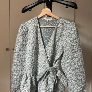 Blommig omlottklänning från View - Supersöt ljusblå omlottklänning med vita blommor från View. Klänningen har långa puffärmar, knyt i midjan och v-ringning. Perfekt för dig som gillar romantiska vibes och vill sticka ut lite extra.