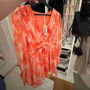 Lovisa Wallin klänning  - Superfin kort omlottklänning från NA-KD i korallorange och vitt blommigt mönster. Klänningen har v-ringning, markerad midja och långa, luftiga ärmar. Tillverkad i återvunnen polyester för en lätt och flowy känsla. Perfekt för sommarens alla tillfällen! (ALDRIG ANVÄND, och säljs inte mer.)💕 Därav priset 