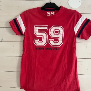 Röd t-shirt med tryck 59 - Sportig röd t-shirt med stort vitt 59-tryck och texten 'Authentic Training Apparel' på bröstet. Ärmarna har vita och marinblå ränder för en cool retrostil. Perfekt för dig som gillar en avslappnad och aktiv look.