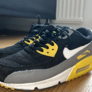 Nike Air Max 90 svart/gul/grå sneaker - Nike Air Max 90 sneakers med svart mesh och skinn, gråa och vita detaljer samt gula inslag på sula och snörningspanel. Klassisk Air Max-dämpning och tydlig logga på hälen. Snygg kontrast mellan färgerna och ikonisk siluett som sticker ut.