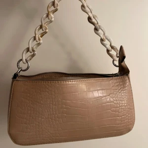 beige axelväska med kedjehandtag - • säljer en beige axelväska i croco-mönstrat skinnimitation med chunky kedjehandtag i vit plast. • väskan har dragkedja upptill och en stilren, avlång form som ger en trendig vibe. • perfekt för dig som gillar detaljer och vill sticka ut lite extra.