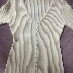 Beige ribbad kofta med v-ringning - Snygg beige ribbad kofta med djup v-ringning och pärlemorknappar längs hela framsidan. Långärmad och figurnära passform, perfekt att styla med jeans eller kjol. Mjuk och stretchig stickad känsla som ger en clean och trendig look.