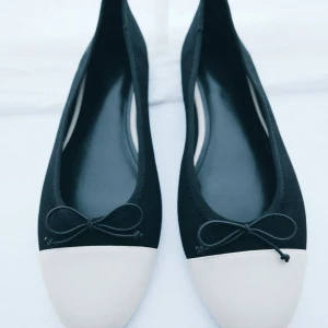 Svarta ballerinaskor med rosett - Stilrena ballerinaskor i svart tyg med vit tå och söt liten rosett framtill. Skorna har rund tå och platt sula, perfekta för en chic och enkel look. Materialet är mjukt och bekvämt, och färgkombon ger en klassisk vibe.