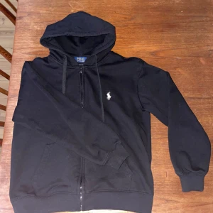 Svart hoodie från Polo Ralph Lauren - Svart hoodie från Polo Ralph Lauren med dragkedja och klassisk logga broderad på bröstet. Tröjan har huva med dragsko och två fickor framtill. Perfekt för en avslappnad och stilren look.